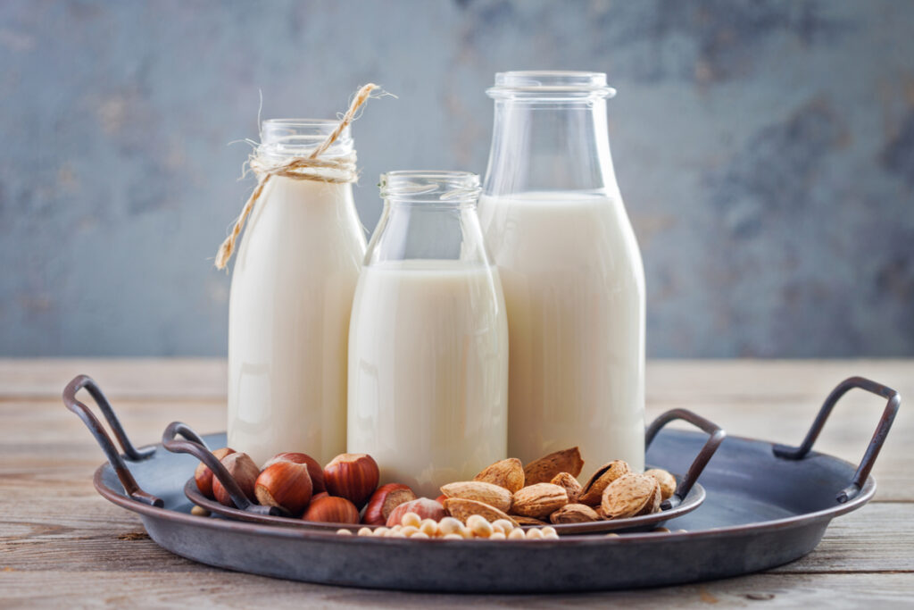 Leite Vegetal Caseiro- Três copos com leites vegetais caseiros: aveia, amêndoas, sobre prato com ingredientes ao redor.